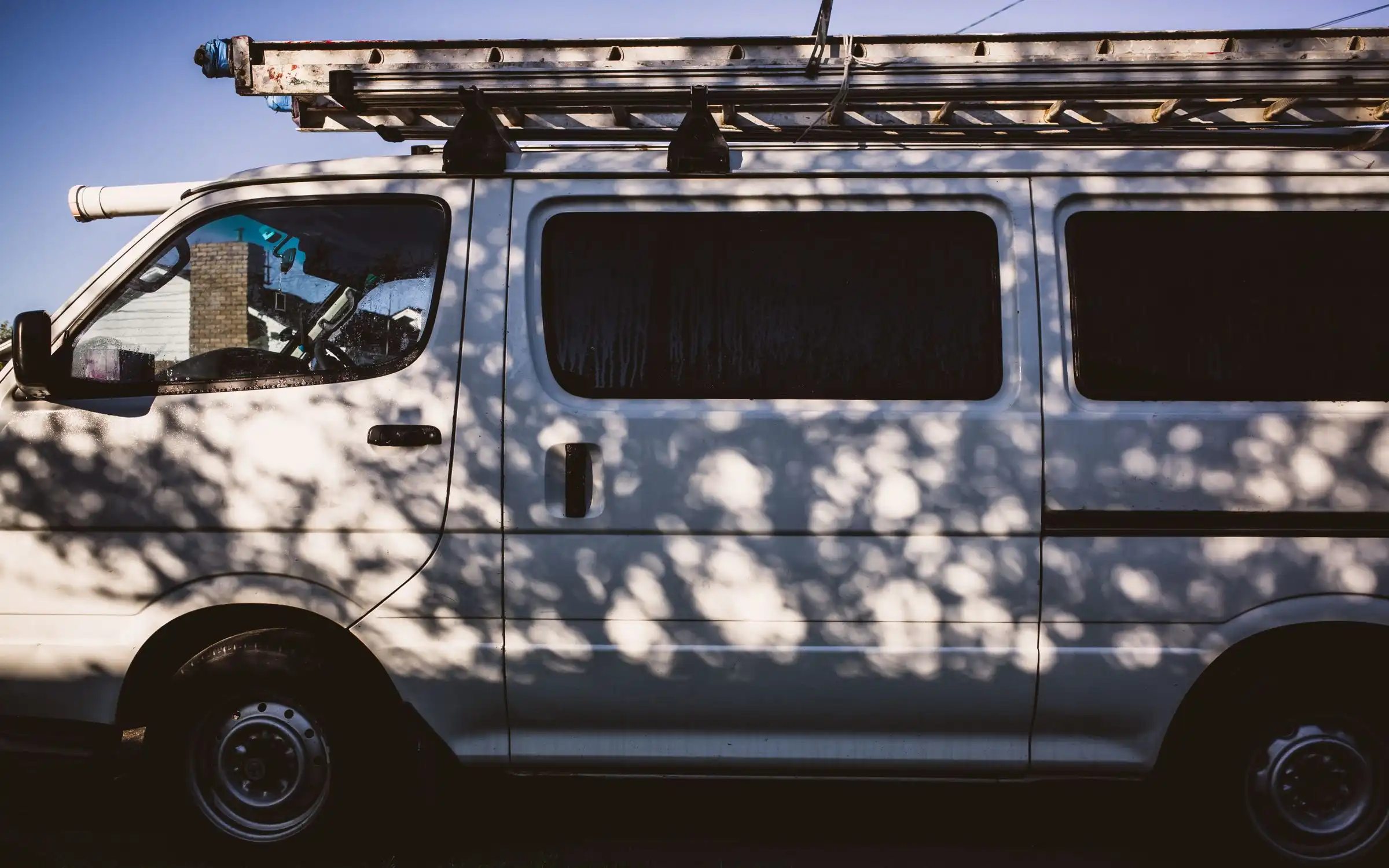 The side of a white work van with a ladder on its roof rack, in the shade of a tree.